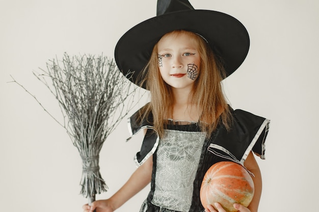 A young girl dressed in black as a witch has a cone-shaped hat on her head. Girl holding a broom and a pumpkin