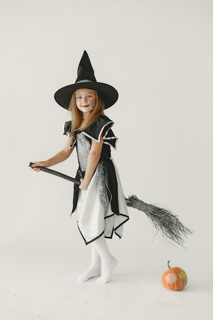 A young girl dressed in black as a witch has a cone-shaped hat on her head. Girl sitting on a broom and have a pumpkin