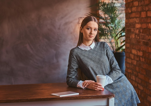 Giovane ragazza vestita con un elegante abito grigio tiene una tazza di caffè da asporto guardando una telecamera mentre si appoggia sul tavolo in una stanza con interni soppalcati.