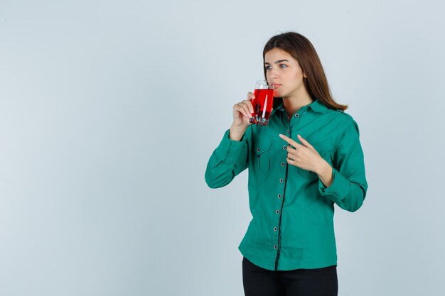 Young girl drinking glass of red liquid, pointing at it with index finger in green blouse, black pants and looking focused. front view.