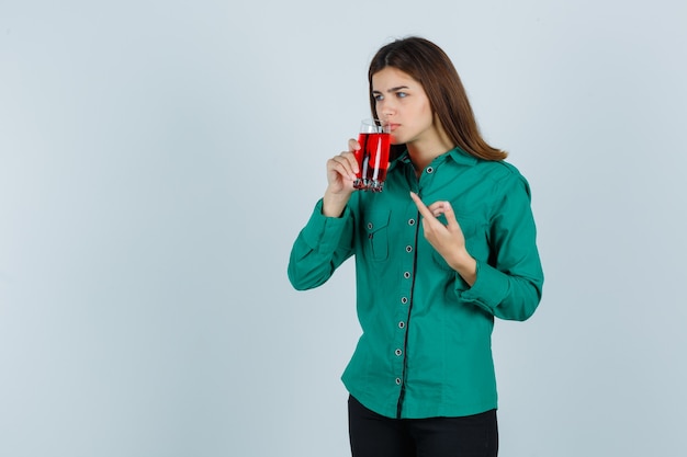 Young girl drinking glass of red liquid, pointing at it with index finger in green blouse, black pants and looking focused. front view.