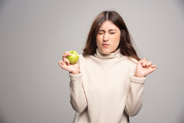 A young girl eating a green apple on a gray wall.