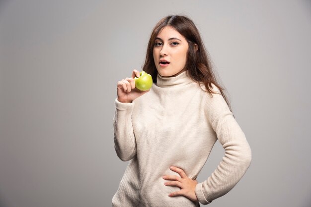 A young girl eating a green apple on a gray wall.