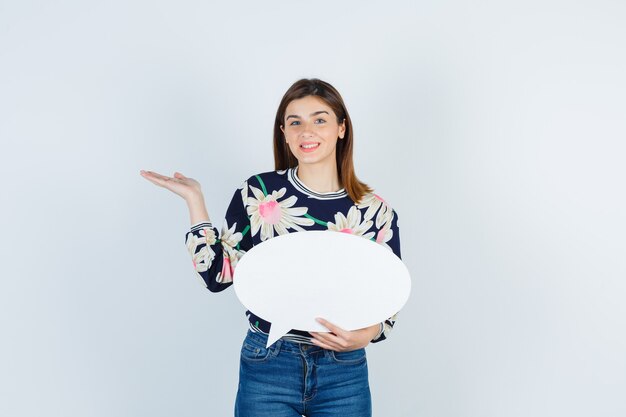 Young girl in floral blouse, jeans spreading palm and looking happy , front view.