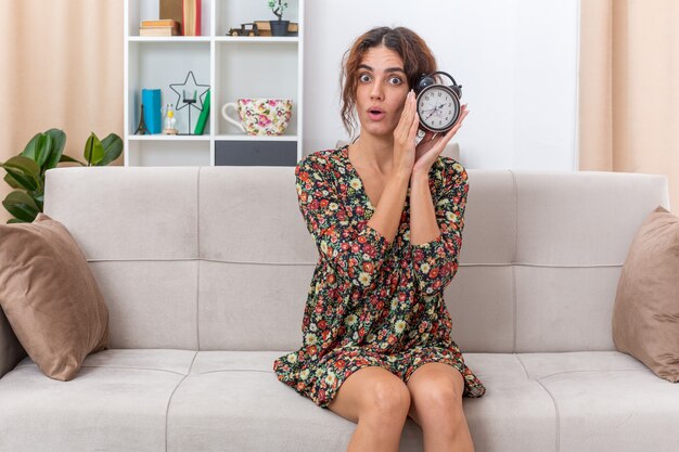 Young girl in floral dress holding alarm clock looking surprised sitting on a couch in light living room