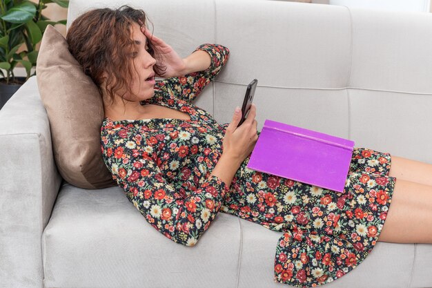 Young girl in floral dress with book and smartphone looking at it confused lying on a couch in light living room