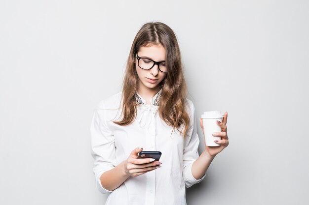 Young girl in glasses dressed up in strict office white t-shirt stands in front of white wall and holds her phone and coffee cup in hands