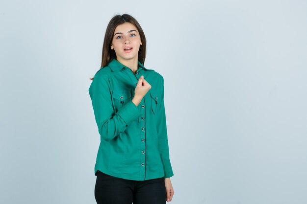 Young girl in green blouse, black pants clenching fist over chest and looking cheery , front view.