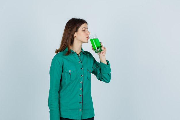 Young girl in green blouse, black pants drinking glass of green liquid and looking focused , front view.