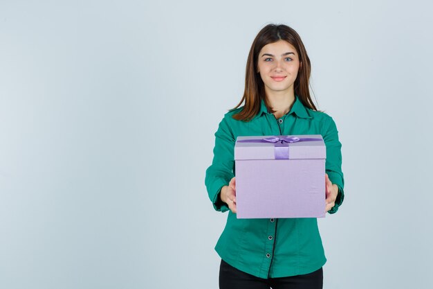 Young girl in green blouse, black pants giving gift box and looking happy , front view.