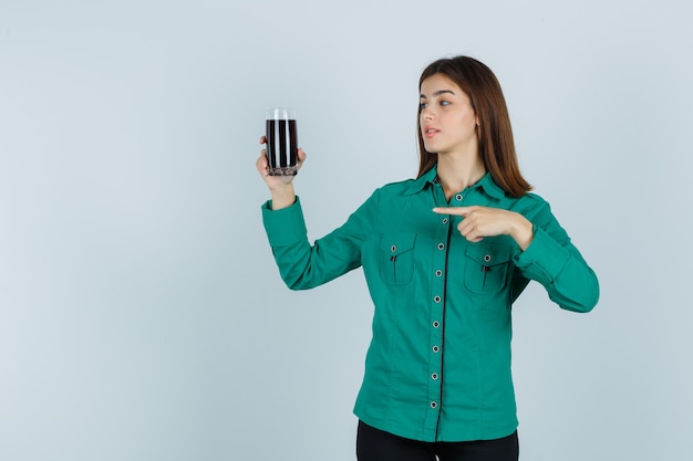 Young girl in green blouse, black pants holding glass of black liquid, pointing at it and looking focused , front view.