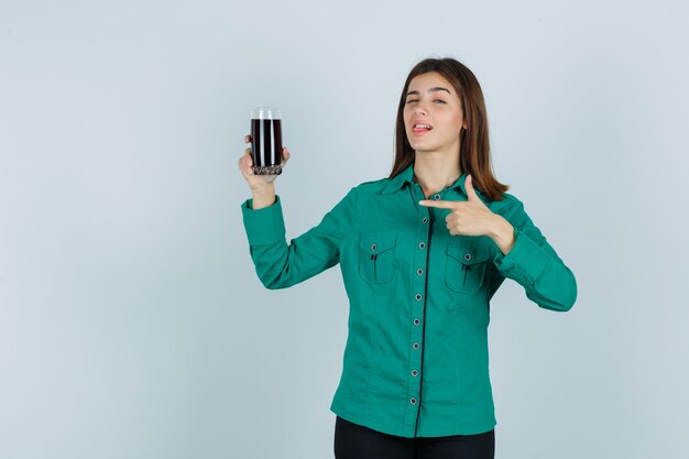 Young girl in green blouse, black pants holding glass of black liquid, pointing at it, winking and looking cute , front view.