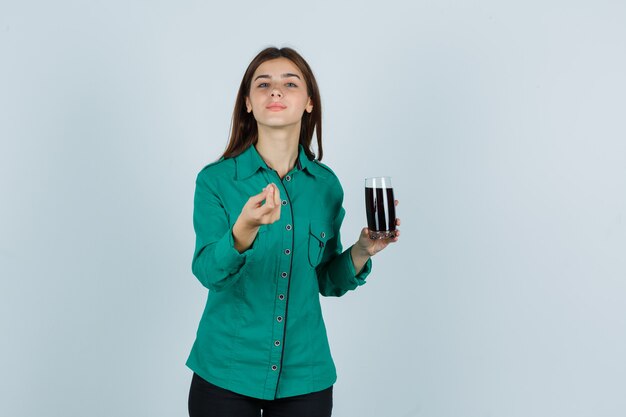 Young girl in green blouse, black pants holding glass of black liquid, showing Italian gesture and looking satisfied , front view.