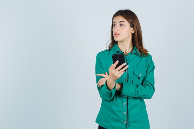 Young girl in green blouse, black pants holding phone, looking away and looking focused , front view.