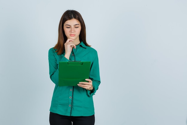 Young girl in green blouse, black pants looking at clipboard, propping chin and looking focused , front view.