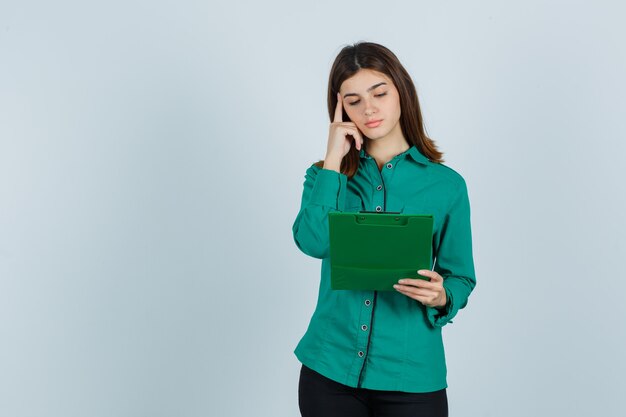 Young girl in green blouse, black pants looking at clipboard, putting index finger on temple and looking pensive , front view.