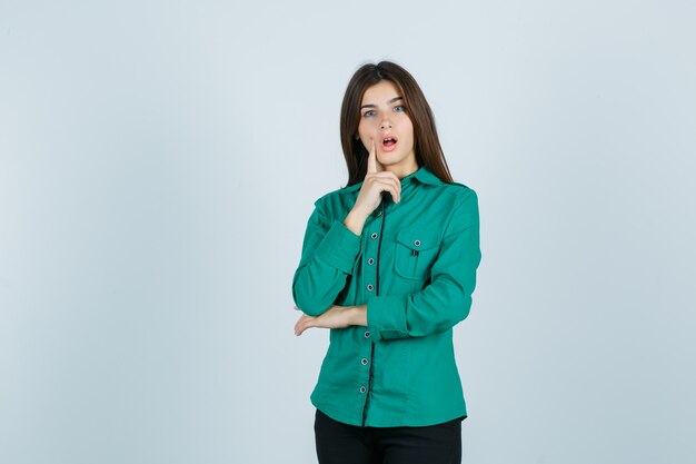 Молодая девушка в зеленой блузке, черных штанах, прикладывая указательный палец ко рту, держа рот широко открытым и выглядя удивленным, вид спереди.