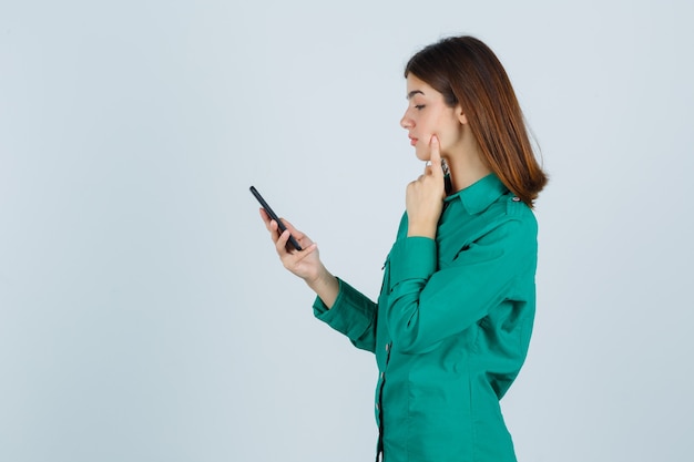 Young girl in green blouse, black pants reading messages on phone, putting index finger on cheek and looking focused , front view.