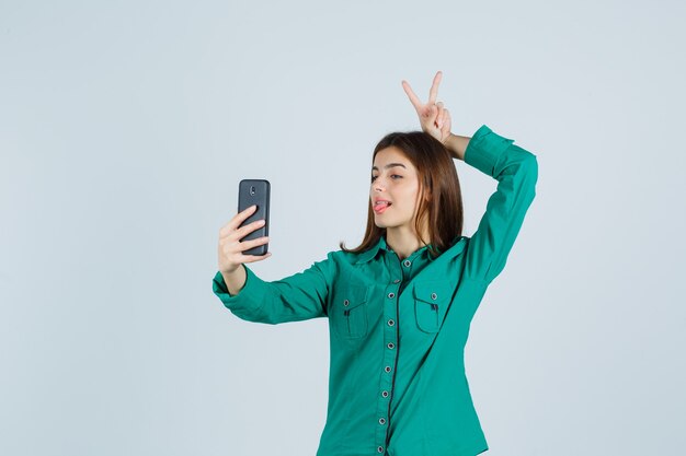 Молодая девушка в зеленой блузке, черных штанах показывает жест мира над головой во время видеозвонка и выглядит удивленным, вид спереди.