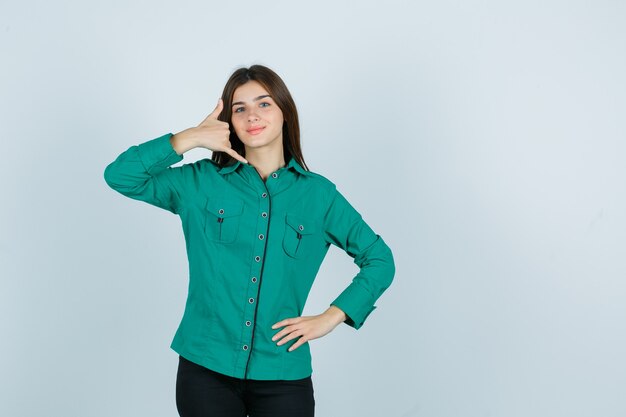 Young girl in green blouse, black pants showing phone gesture, holding hand on hip and looking sanguine , front view.