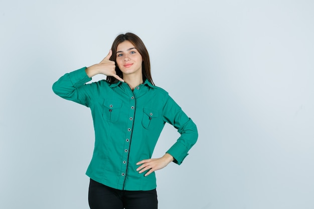 Молодая девушка в зеленой блузке, черных штанах показывает жест телефона, держит руку на бедре и выглядит оптимистично, вид спереди.