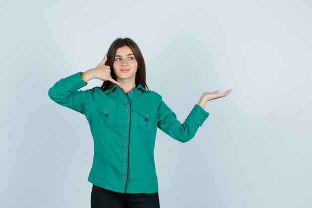 Молодая девушка в зеленой блузке, черных штанах показывает жест телефона, разводит ладонь и выглядит оптимистично, вид спереди.