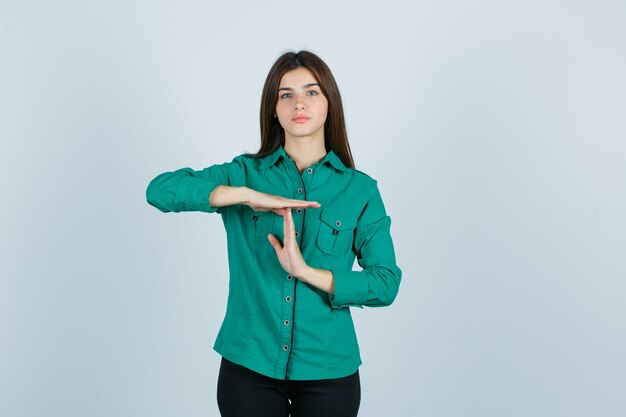 Young girl in green blouse, black pants showing time-break gesture and looking serious , front view.