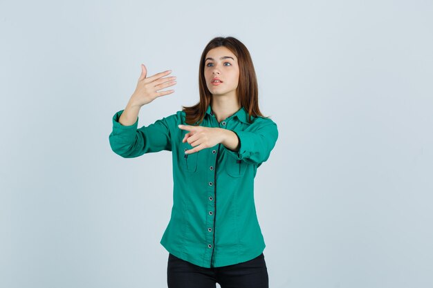 Young girl in green blouse, black pants stretching hand as holding something imaginary, showing rock n roll gesture and looking focused , front view.