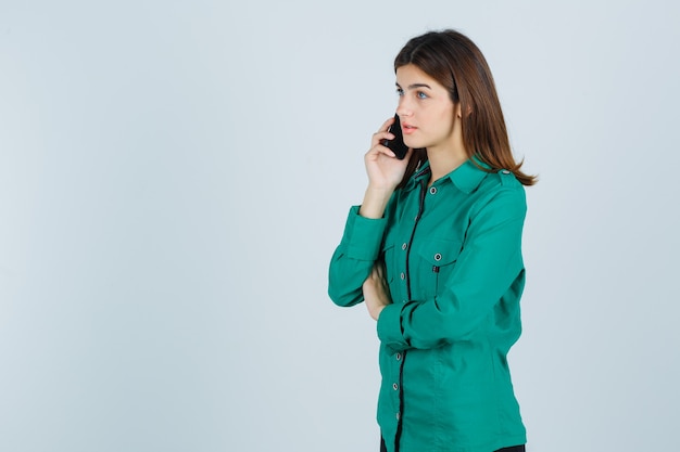 Young girl in green blouse, black pants talking to phone and looking focused , front view.