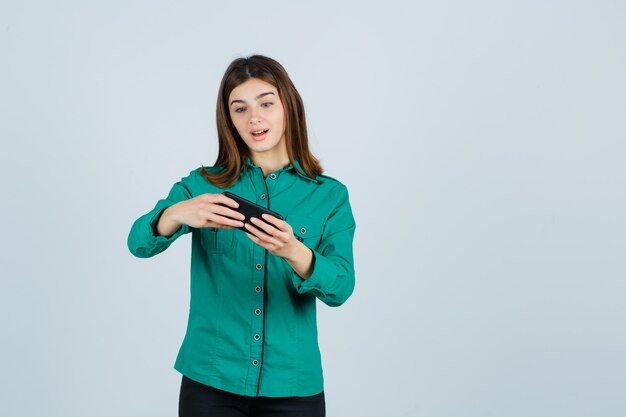 Молодая девушка в зеленой блузке, черных штанах смотрит видео по телефону и удивленно смотрит, вид спереди.
