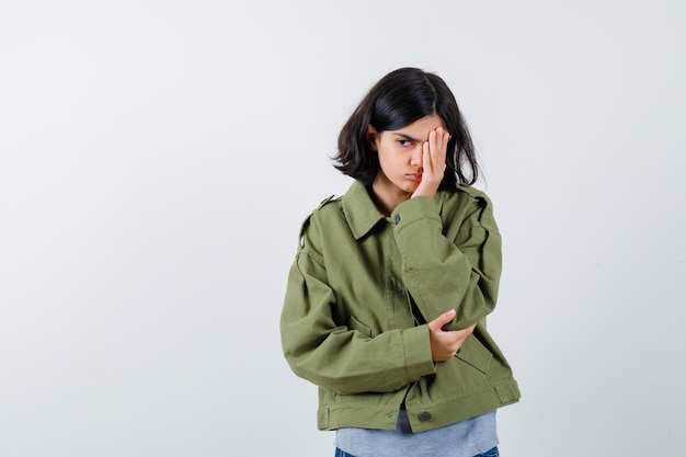 Молодая девушка в сером свитере, куртке цвета хаки, джинсовых штанах, прикрывающих глаз рукой, держащей руку на локте и задумчивой, вид спереди.