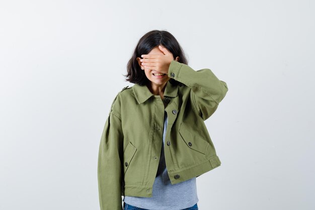 Молодая девушка в сером свитере, куртке цвета хаки, джинсовых брюках, закрывающих глаза рукой и выглядящих мило, вид спереди.