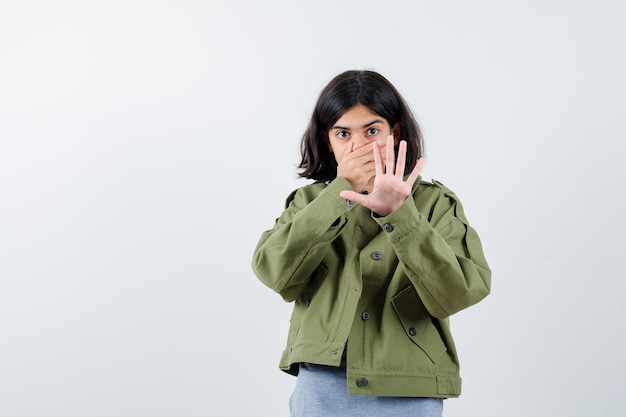 Giovane ragazza in maglione grigio, giacca color kaki, pantalone di jeans che copre la bocca, mostra il segnale di stop e sembra sorpresa, vista frontale.