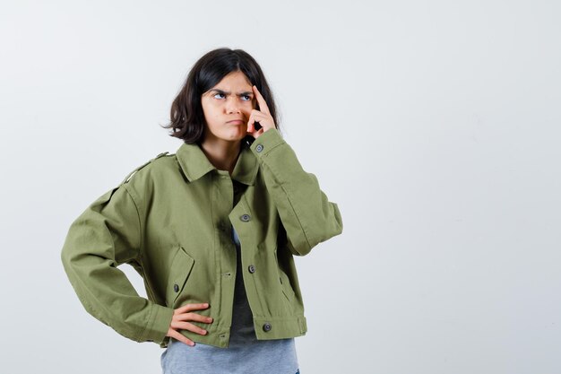 Young girl in grey sweater, khaki jacket, jean pant holding hand on waist while scratching head and looking pensive , front view.