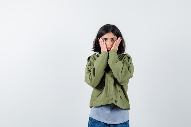 Young girl in grey sweater, khaki jacket, jean pant holding hands on cheeks and looking surprised , front view.