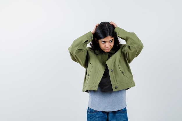 Giovane ragazza in maglione grigio, giacca kaki, pantaloni di jeans che si tengono per mano sulla testa, distogliendo lo sguardo e guardando pensieroso, vista frontale.