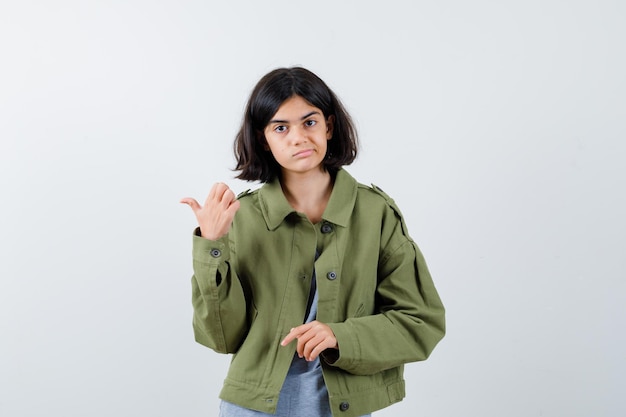 Young girl in grey sweater, khaki jacket, jean pant pointing left and looking serious , front view.
