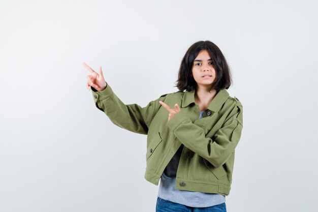 Young girl in grey sweater, khaki jacket, jean pant pointing left with index fingers and looking serious , front view.