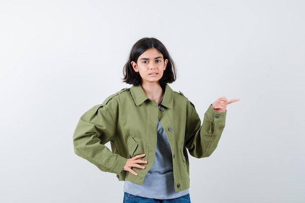 Молодая девушка в сером свитере, куртке цвета хаки, джинсовых штанах, указывающих вправо, держа руку на талии и серьезно выглядящих, вид спереди.