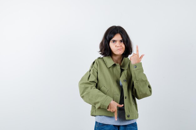 Young girl in grey sweater, khaki jacket, jean pant pointing up and down and looking serious , front view.
