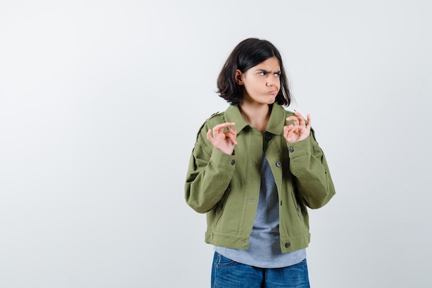 Young girl in grey sweater, khaki jacket, jean pant raising palms in surrender gesture and looking furious , front view.