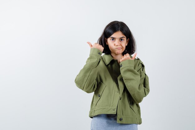 회색 스웨터, 카키색 재킷, 진 바지를 입은 어린 소녀가 양손으로 엄지손가락을 치켜들고 행복해 보이는 앞모습을 보여줍니다.