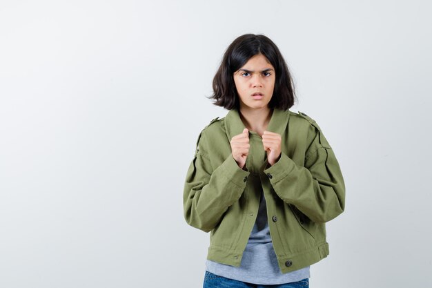 Young girl in grey sweater, khaki jacket, jean pant standing in boxer pose and looking serious , front view.
