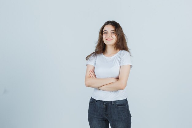 Young girl holding arms folded in t-shirt, jeans and looking pleased . front view.