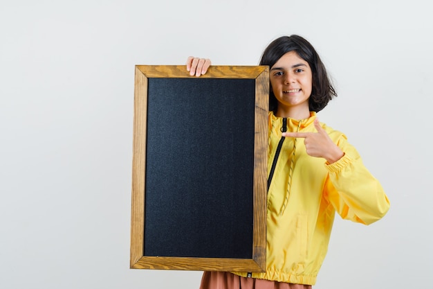 Young girl holding blackboard, pointing to it with index finger in yellow bomber jacket and looking happy.