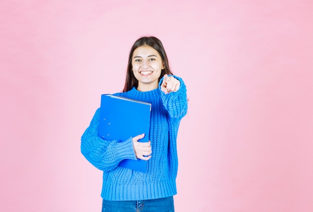 青いフォルダーを保持し、ピンクの正面を指している若い女の子。