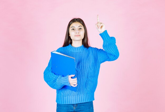 young girl holding a blue folder and pointing up on pink.