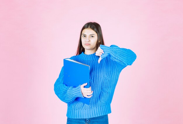 young girl holding a blue folder and showing a thumb down on pink.