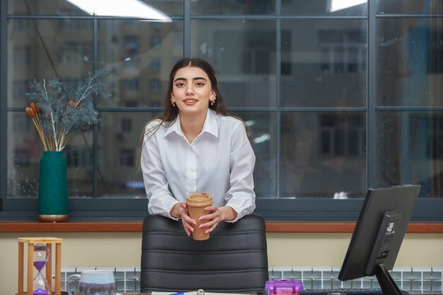 Молодая девушка держит чашку кофе и опирается на офисный стул