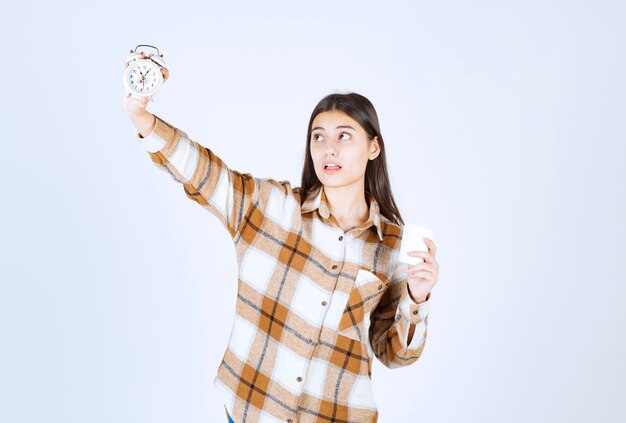 Young girl holding cup of tea and looking at time on white wall. 